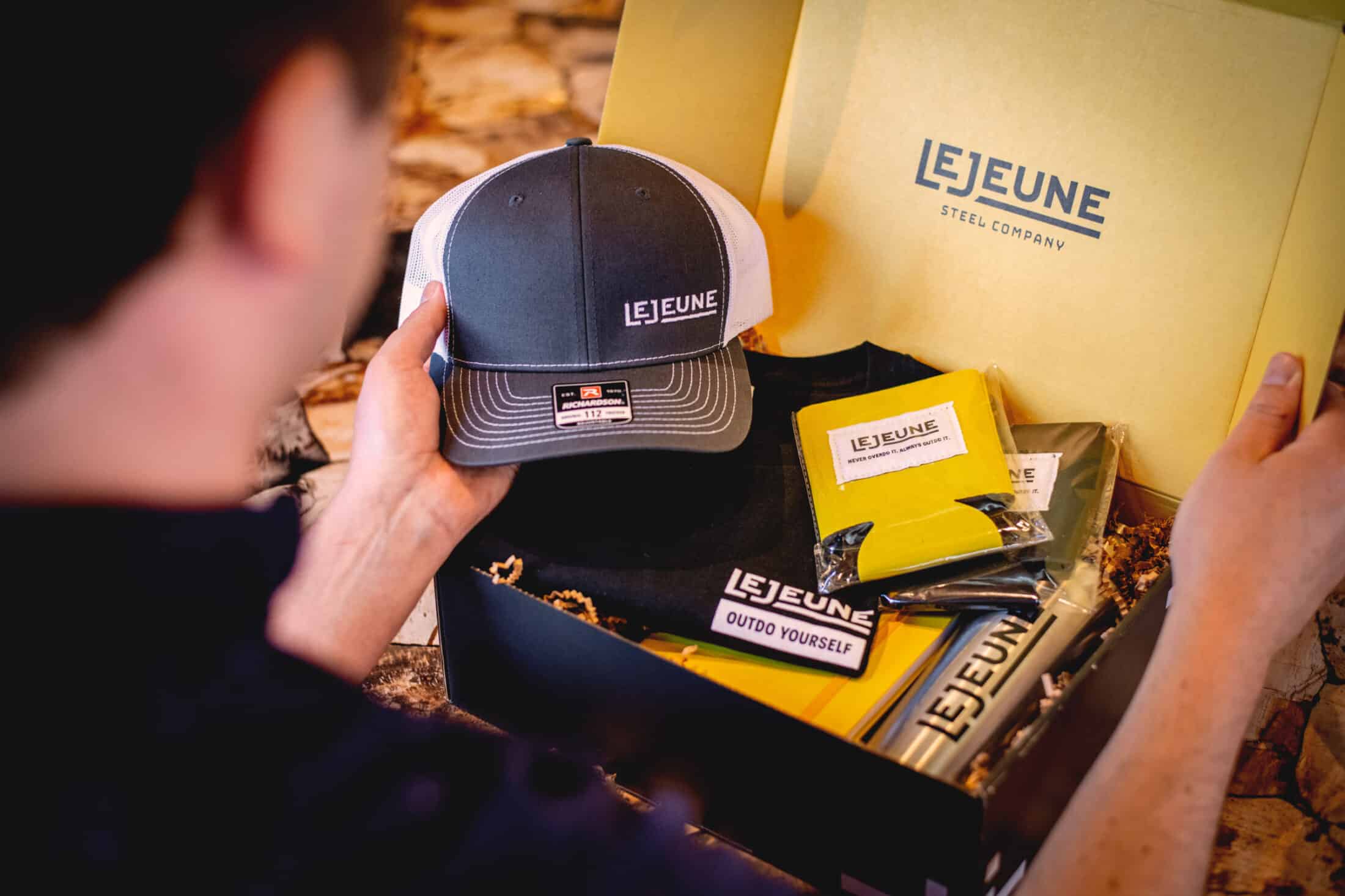 Man opening a gift box of company-branded items including a hat, water bottle, shirt and koozie.
