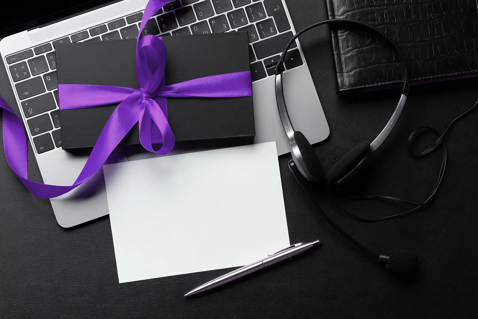 Computer, headphones, paper, pen and a gift box wrapped with purple ribbon on a desk